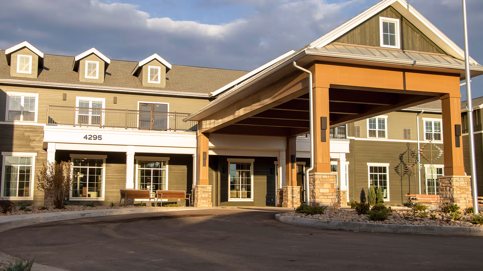 Exterior view of a senior living facility building with a covered entrance supported by stone and wood pillars, multiple windows, and a driveway in front. The building has a green and beige color scheme with a sloped roof and dormer windows.