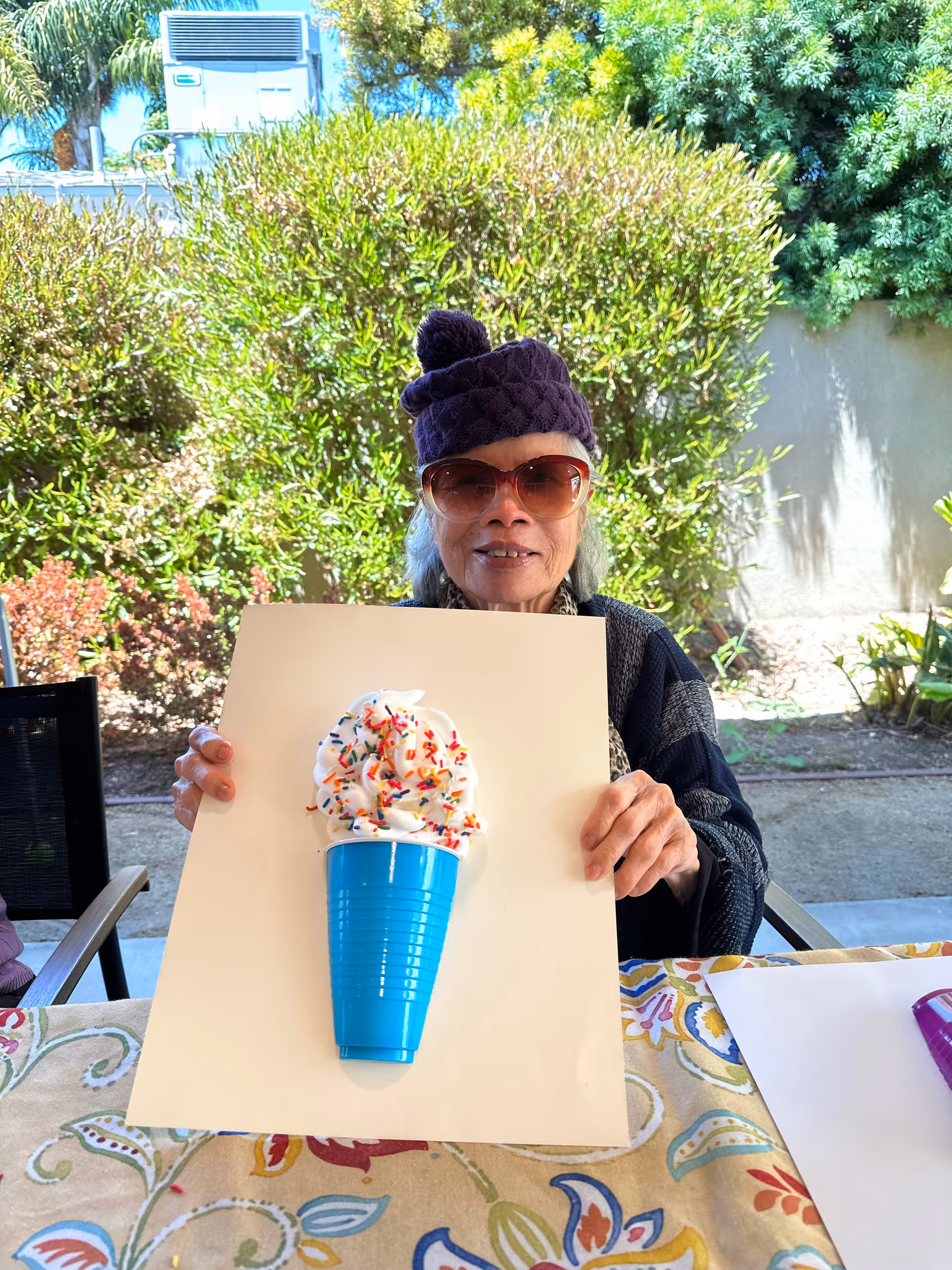 An elderly woman wearing sunglasses and a purple knit hat is sitting outdoors at a table with a colorful tablecloth. She is holding up a white board with a blue plastic cup attached to it, topped with whipped cream and colorful sprinkles, resembling an ice cream cone. Green bushes and trees are visible in the background.