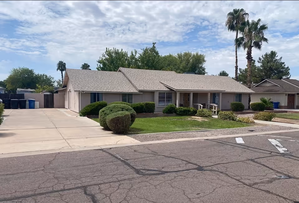 Single-story residential building with a light gray roof and beige exterior walls, surrounded by trimmed bushes and a green lawn. There are palm trees and other trees in the background under a partly cloudy sky. The building has a driveway on the left side and a sidewalk in front.