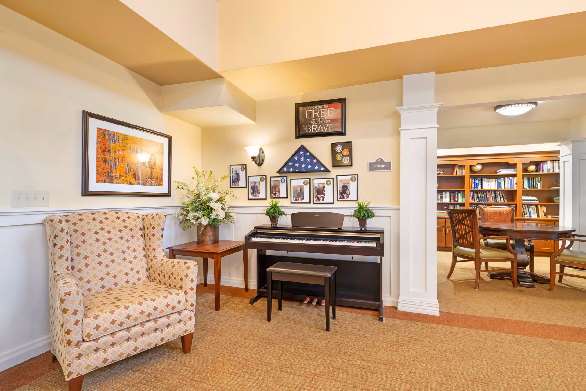 Cozy senior living common area with an upholstered armchair, side table, piano under framed photos and a view into a library with bookshelves and a table.