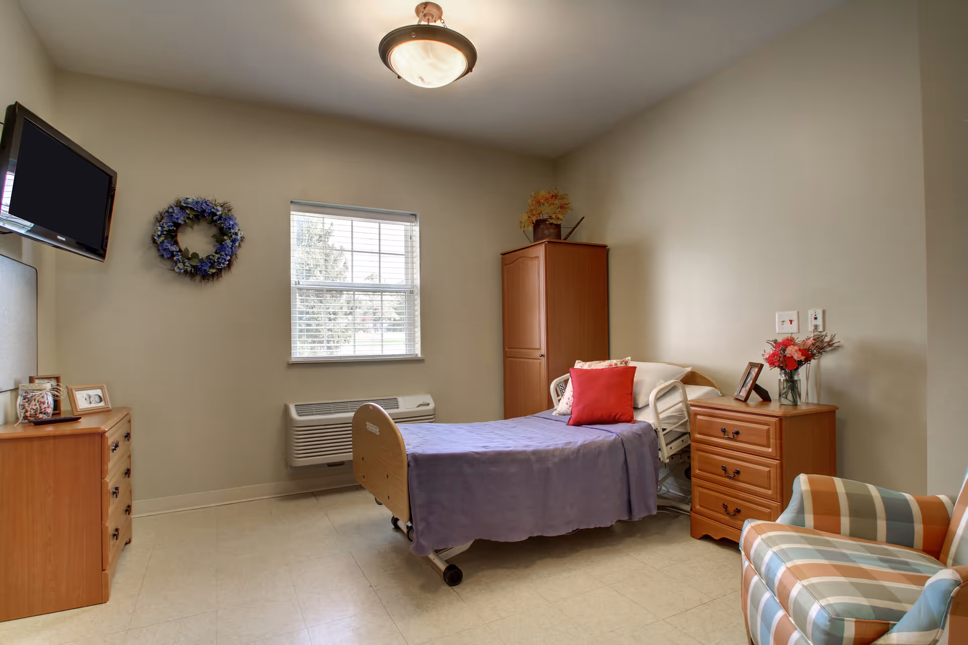 A senior care facility bedroom with a single bed covered in a purple blanket and a red pillow. The room has a wooden dresser with framed photos and a TV mounted on the wall. There is a window with blinds, a wooden wardrobe, a nightstand with a vase of flowers and a framed photo, and a striped armchair in the corner.