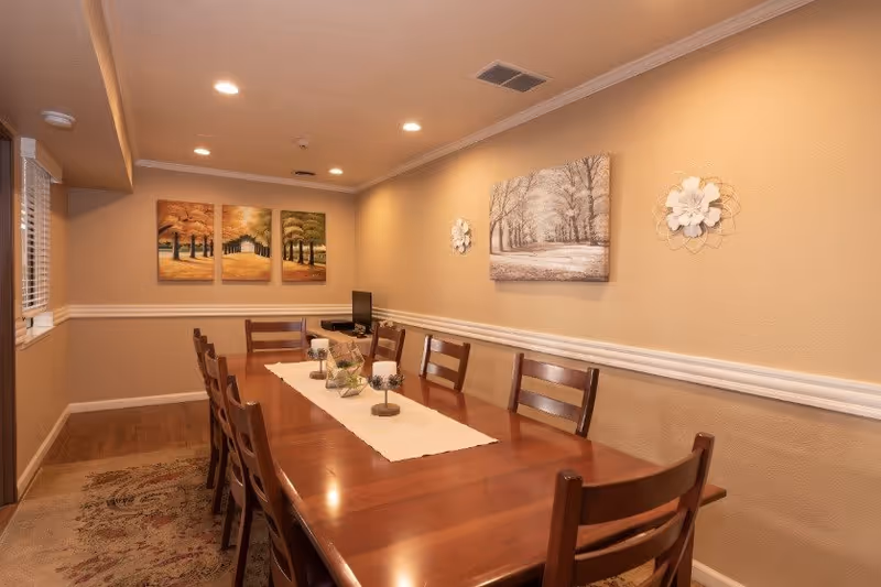 A dining room with a long wooden table surrounded by wooden chairs. The table has a white runner with decorative candles and small plants. The walls are beige with white trim and decorated with three-panel artwork depicting autumn trees and a single large painting of a winter scene, along with two white floral wall decorations. There is a window with blinds on the left side and recessed ceiling lights.