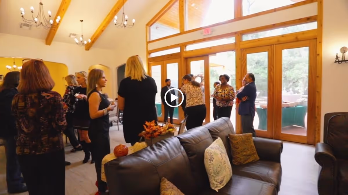 A group of people socializing in a spacious living room with high ceilings and wooden beams. The room features large glass doors leading outside, leather sofas with decorative pillows, and warm lighting from chandeliers.