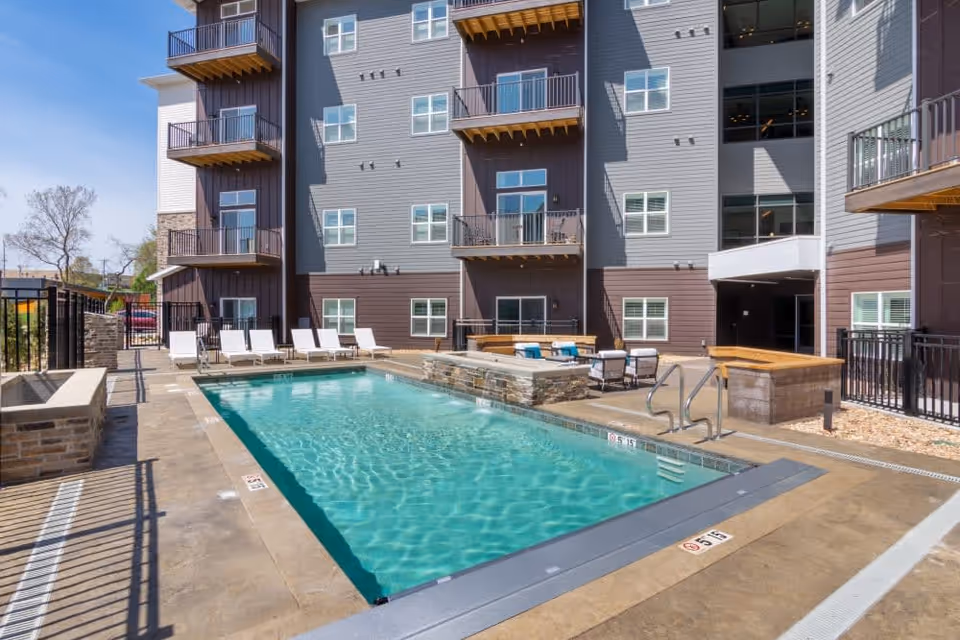 Outdoor swimming pool area at Turners Rock facility with lounge chairs lined up along one side, surrounded by a multi-story building with balconies and windows under a clear blue sky.