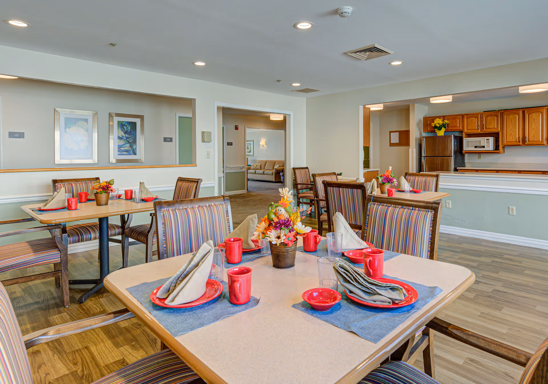 A dining room in a senior living facility with tables set for meals. Each table has red plates, bowls, cups, and folded napkins on blue placemats. There are striped cushioned chairs around the tables. The room has light-colored walls, framed artwork, and a kitchen area with wooden cabinets and stainless steel appliances in the background.