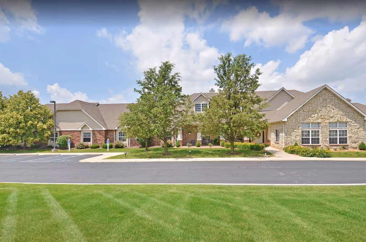 Exterior view of Muncie Estates Senior Living facility showing a single-story building with a combination of brick and stone facade, surrounded by green trees and a well-maintained lawn under a partly cloudy sky.