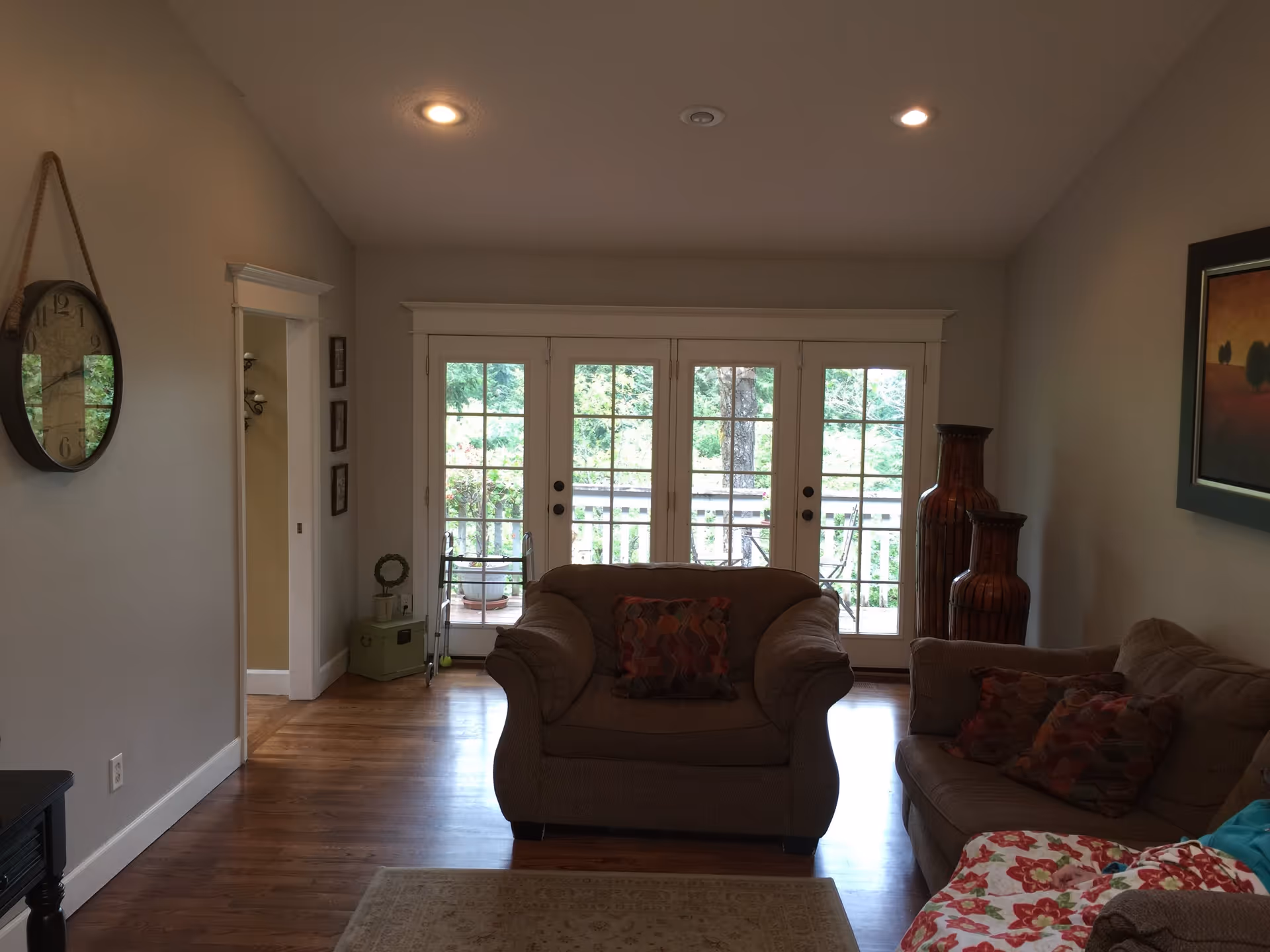 A cozy living room with wooden flooring, a large armchair and a sofa with patterned cushions, a round wall clock hanging on the left wall, two large decorative vases on the right, and a set of French doors leading to an outdoor area with greenery visible outside.