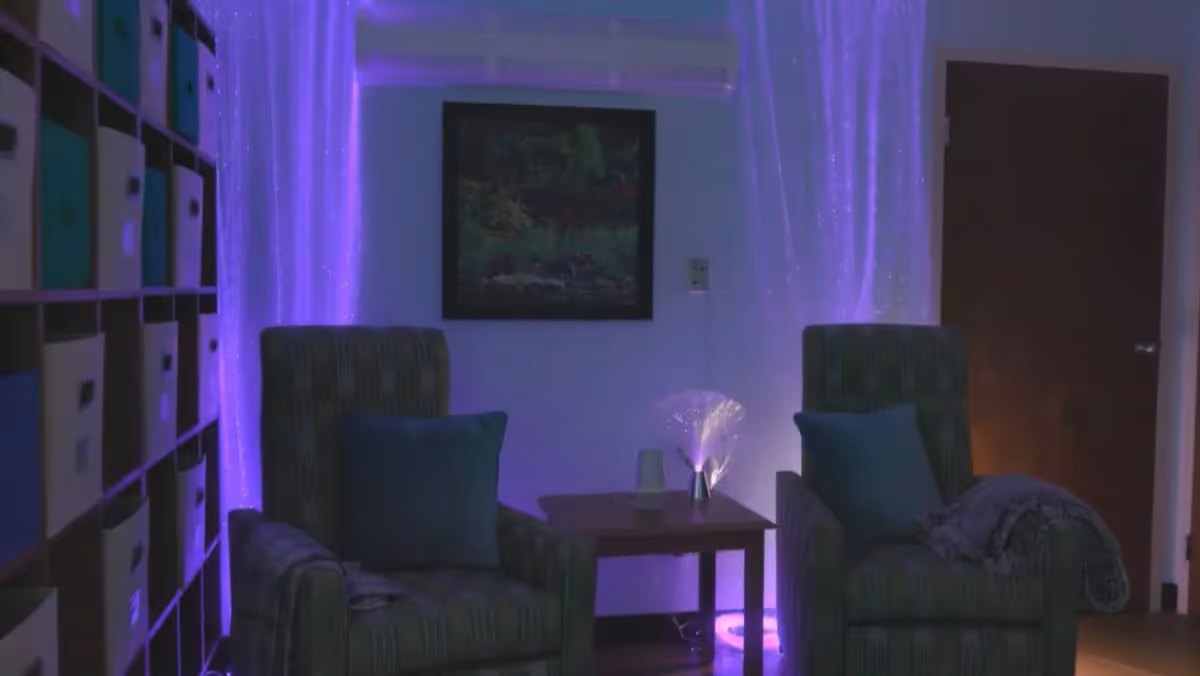 A dimly lit room with two cushioned armchairs facing forward, each with a pillow and a blanket. Between the chairs is a small wooden table with a glowing fiber optic lamp and a cup. Behind the chairs, there is a wall with a framed picture and purple ambient lighting creating a calming atmosphere. To the left, there is a shelving unit with multiple storage bins.