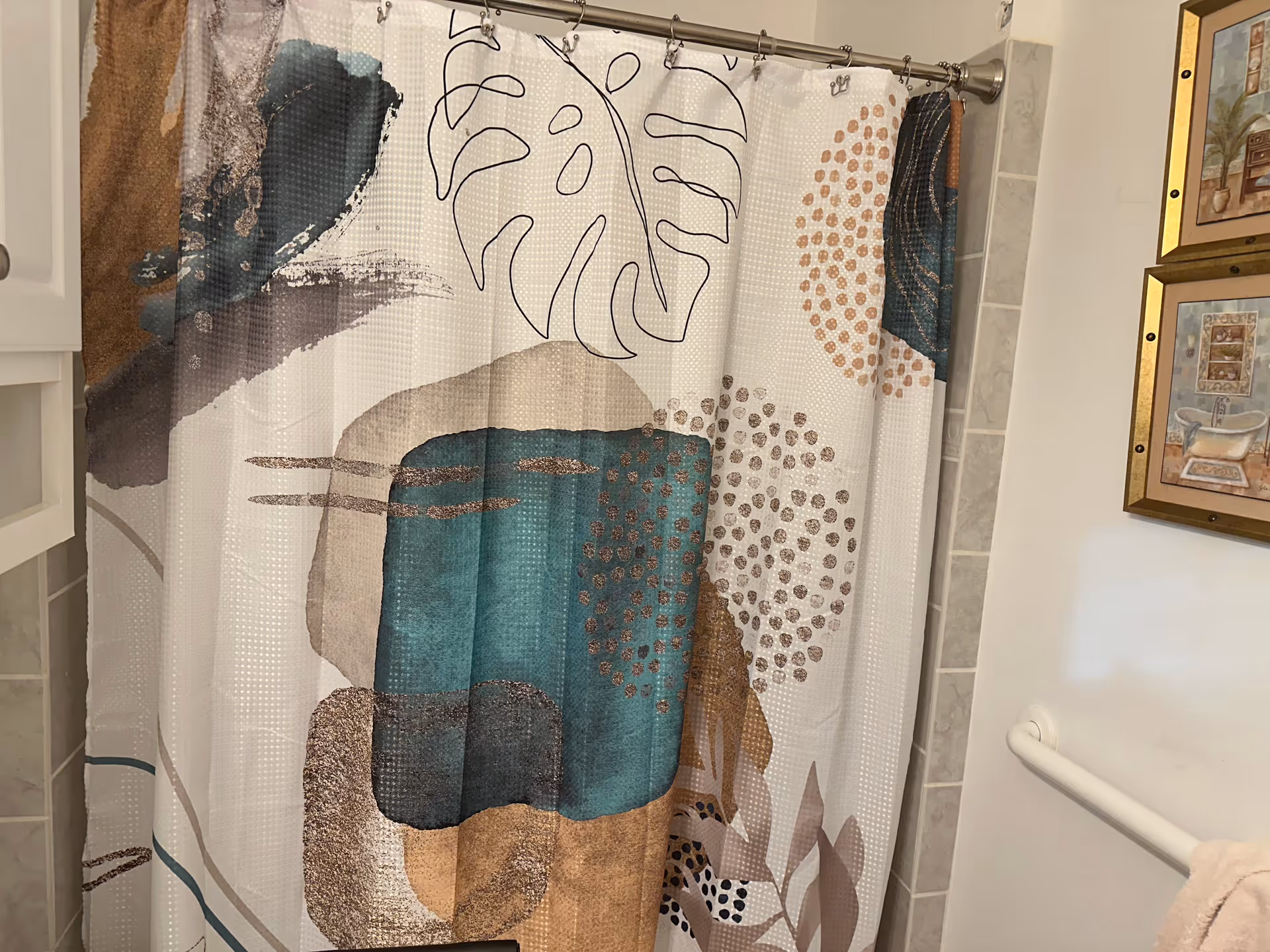 A bathroom with a decorative shower curtain featuring abstract shapes and leaf patterns in teal, brown, and beige colors. There is a white grab bar on the wall and two framed pictures with bathroom-themed artwork hanging above it.