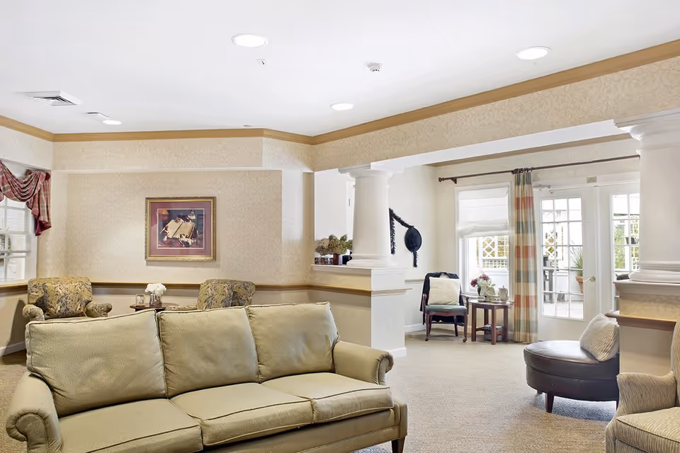 A bright and spacious living room area in a senior living facility with beige walls and carpet. The room features a light green sofa in the foreground, two patterned armchairs with a small round table between them, a framed artwork on the wall, and a separate seating area with a chair, small table, and large windows with curtains. White columns and a glass door leading outside are also visible.