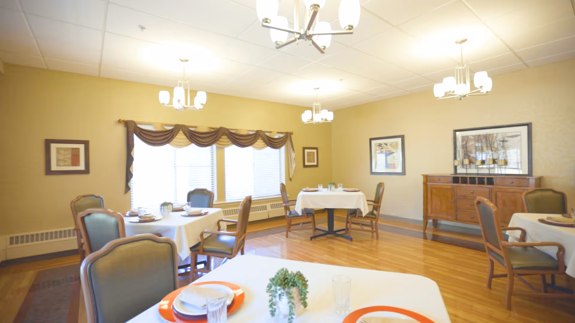 A bright dining room with several round tables covered with white tablecloths, each set with plates, glasses, and napkins. The room has wooden floors, beige walls, and large windows with brown valances. There are green cushioned chairs around the tables, framed artwork on the walls, and a wooden sideboard with decorative items. Ceiling lights provide illumination.