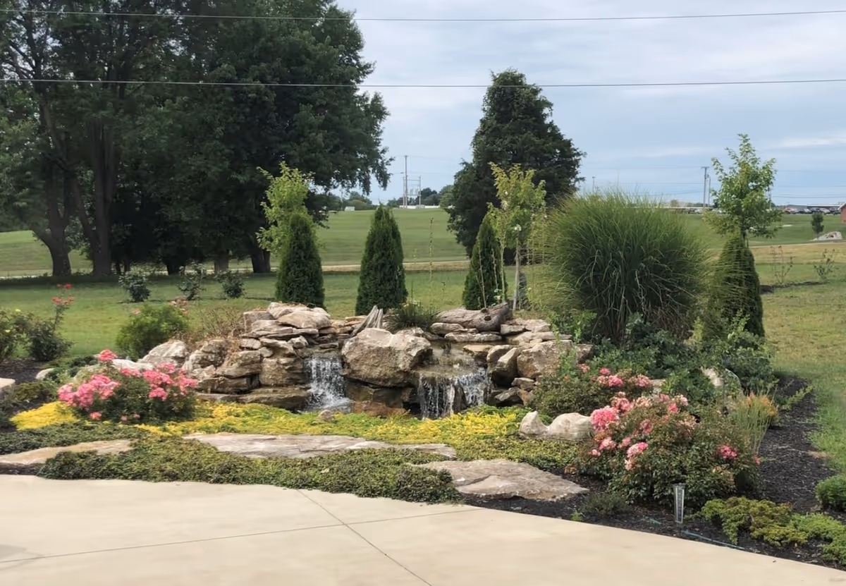 Landscaped outdoor rock waterfall and pond surrounded by flowering shrubs and trees with an open lawn beyond.