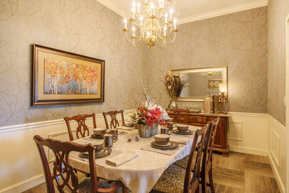 A formal dining room with a table set for six, wooden chairs, a chandelier, framed artwork, and a sideboard with a mirror.