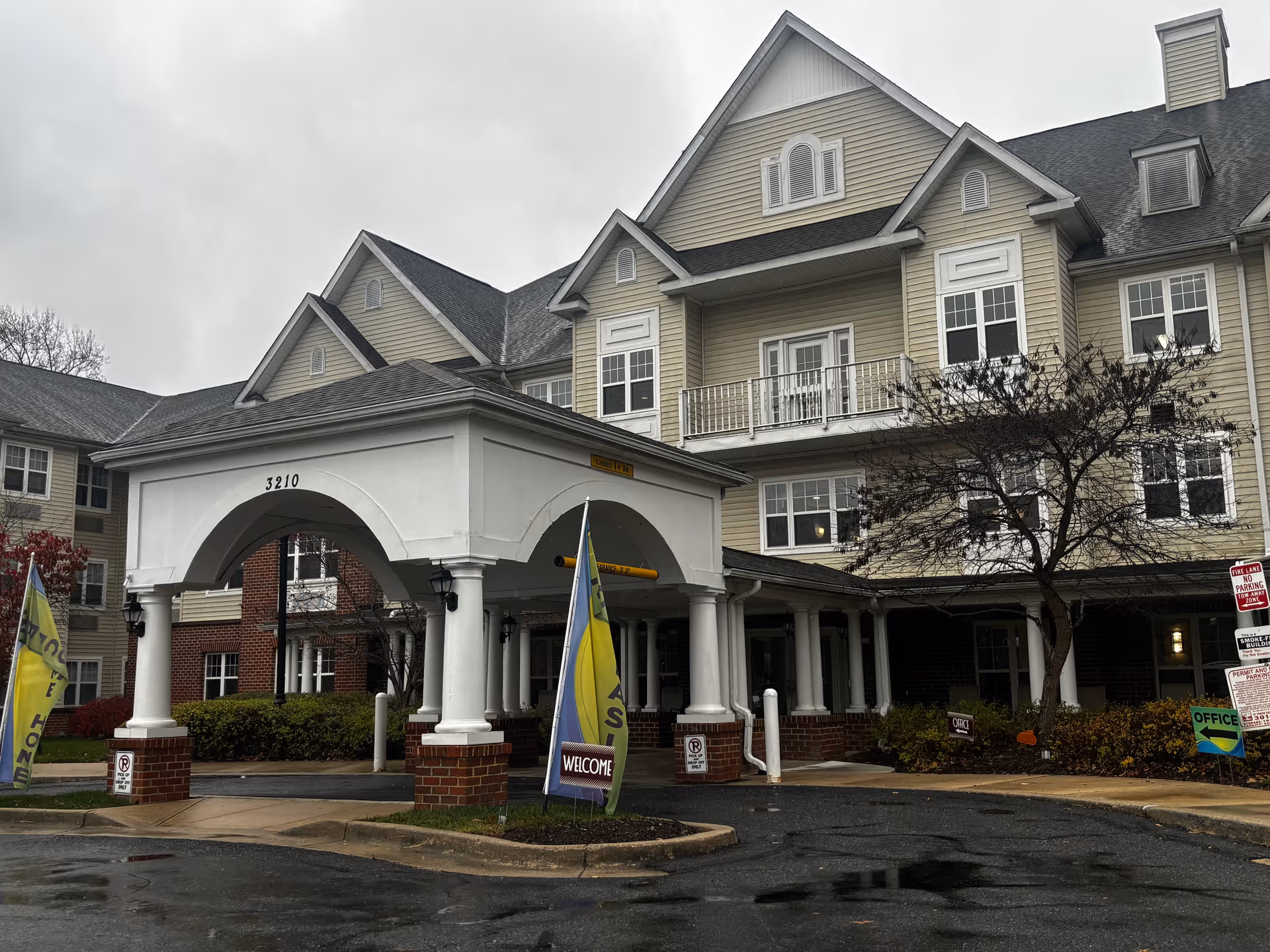 Exterior view of a senior living facility named Grace House on a cloudy day. The building has beige siding with white trim, multiple windows, and a covered entrance with white columns and brick bases. There are flags and signs near the entrance, including one that says 'WELCOME' and another indicating the office location. The ground is wet from recent rain.