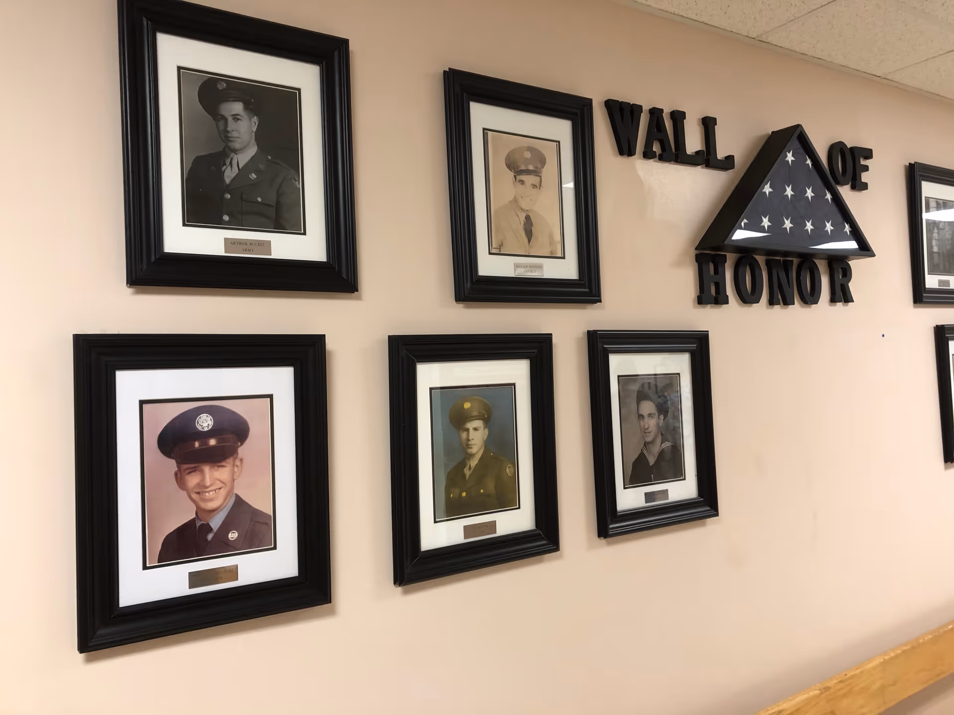 A wall display titled 'Wall of Honor' featuring six framed photographs of military veterans in uniform, arranged around a folded American flag encased in a triangular shadow box.