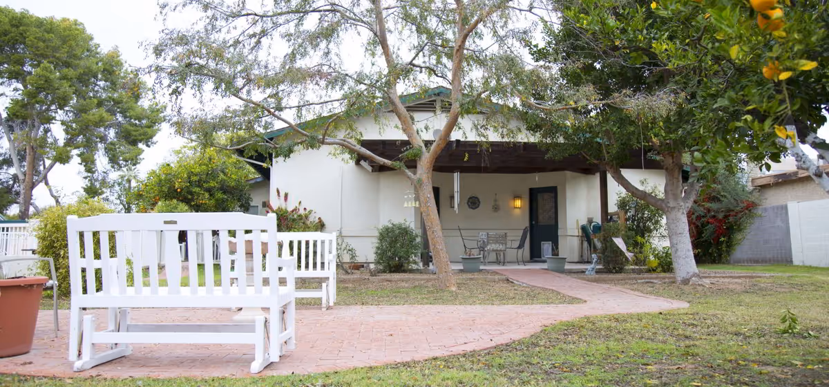 Outdoor patio area of a senior care home with white wooden benches, a brick pathway, trees, and a covered porch with chairs and a table.