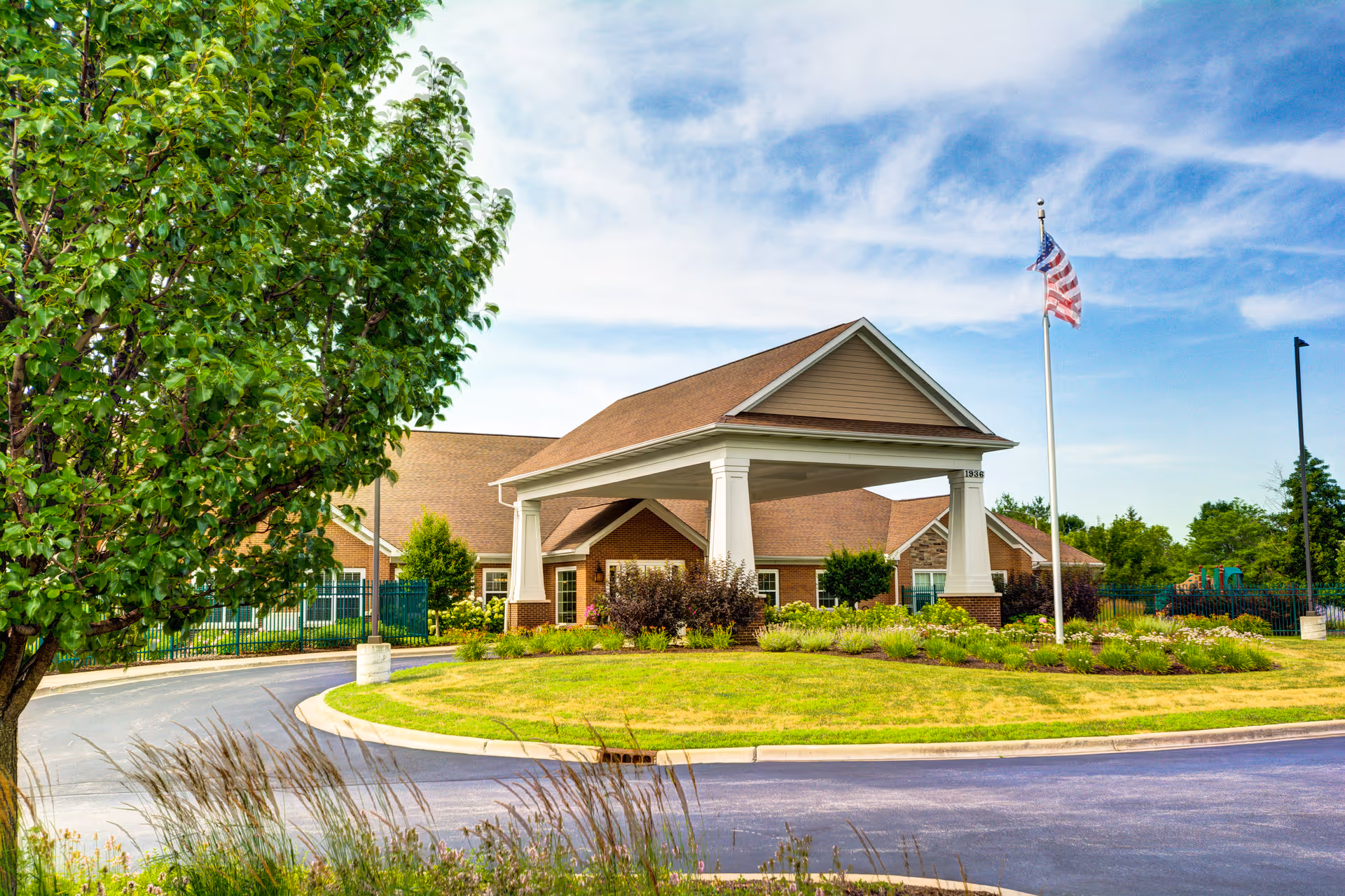 Exterior view of The Auberge at Naperville senior living facility showing a covered entrance with white pillars, a manicured lawn, an American flag on a flagpole, and a tree on the left side under a partly cloudy sky.