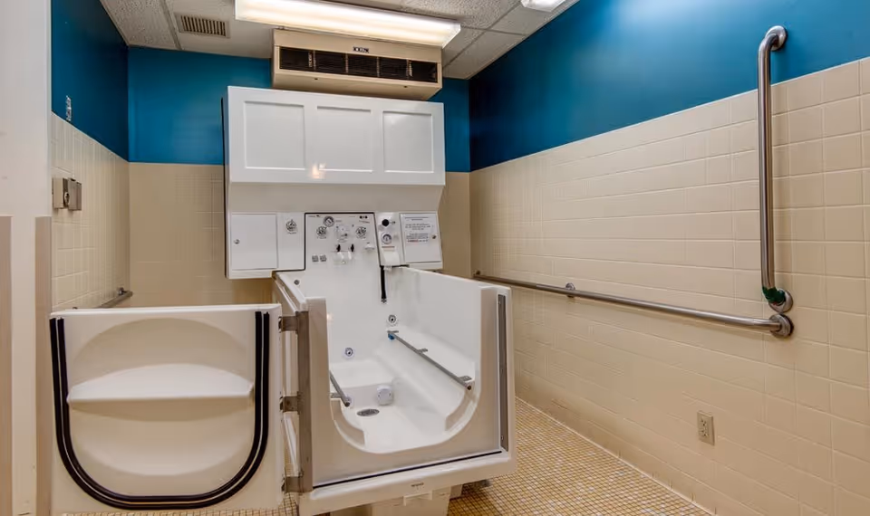 A spacious bathroom with beige tiled walls and floor, featuring a large walk-in bathtub with an open door and built-in handrails. The upper walls are painted teal, and there are metal grab bars installed on the walls for safety.