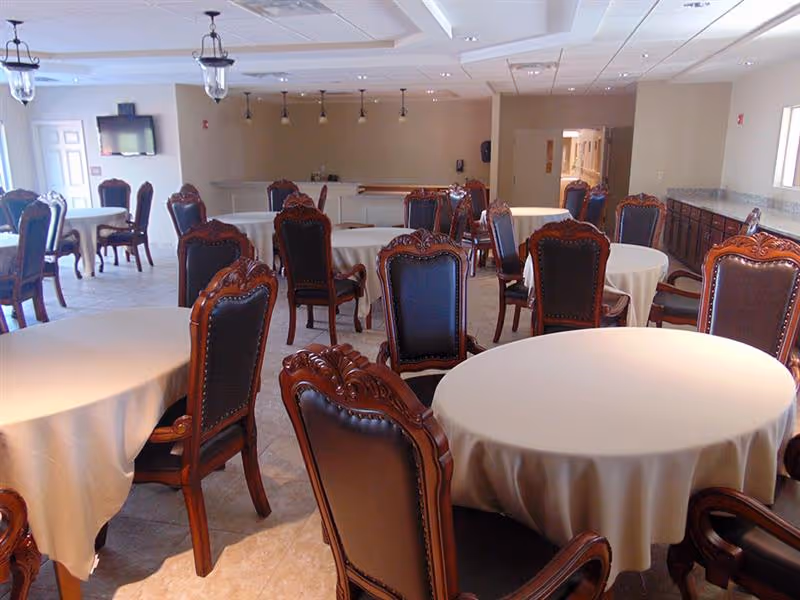 A dining room with multiple round tables covered with white tablecloths and surrounded by ornate wooden chairs with dark leather upholstery. The room has tiled floors, beige walls, and ceiling lights. There is a hallway visible in the background and a wall-mounted television on the left side.
