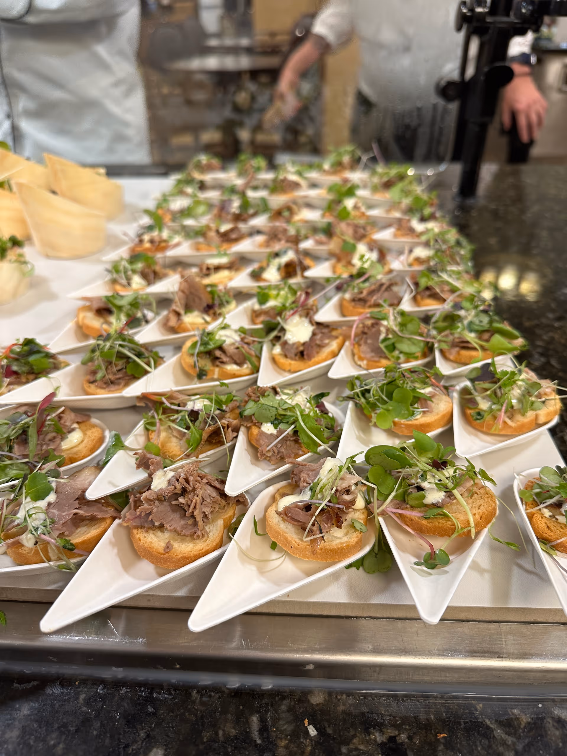 Rows of bite-sized crostini appetizers plated in individual white serving spoons on a counter with kitchen staff visible behind a glass.