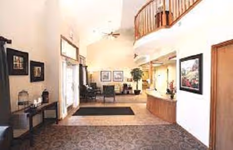 Interior view of a senior living facility lobby with a high ceiling, carpeted floor, seating area with chairs, framed artwork on the walls, a reception desk on the right, and a staircase with wooden railing leading to an upper floor.