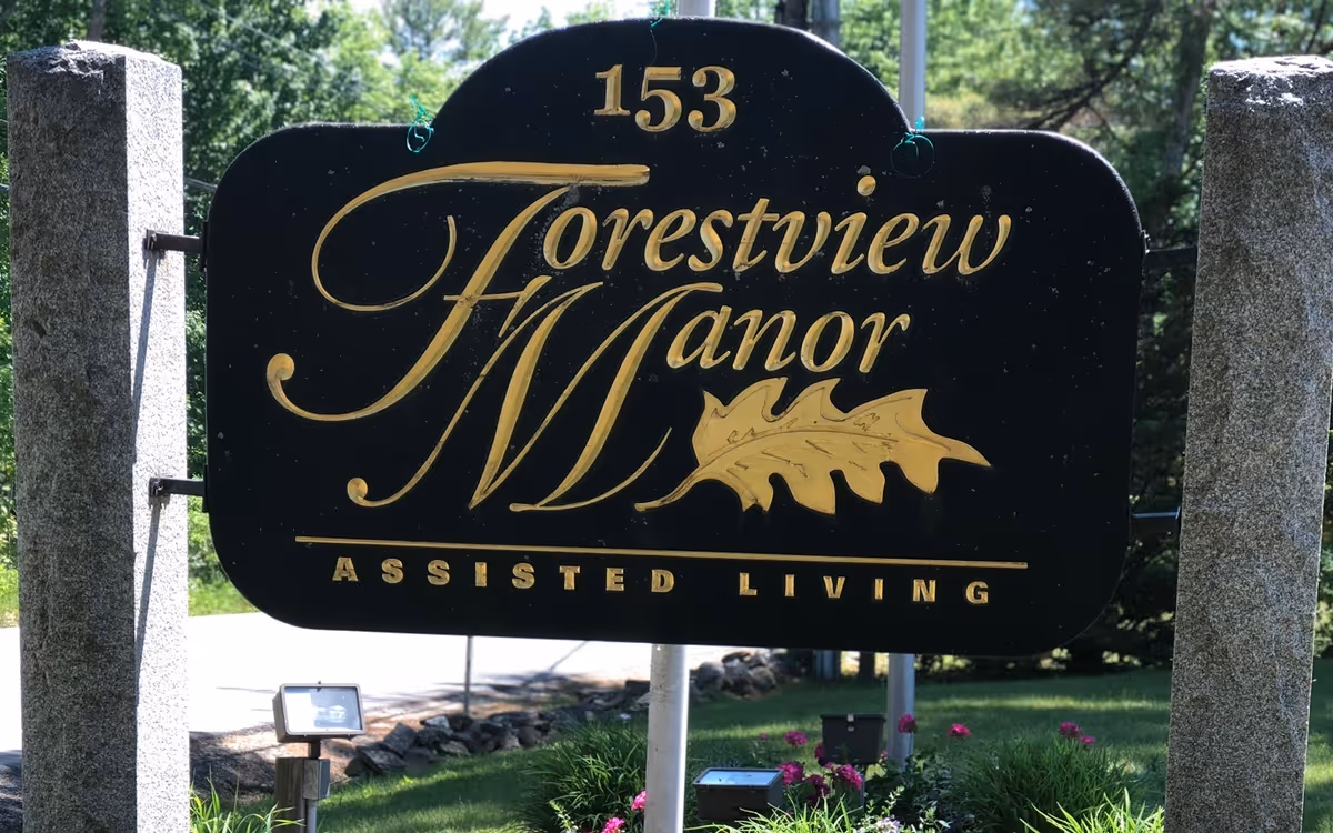 A black and gold sign for Forestview Manor Assisted Living, mounted between two stone pillars with greenery and flowers in the background.