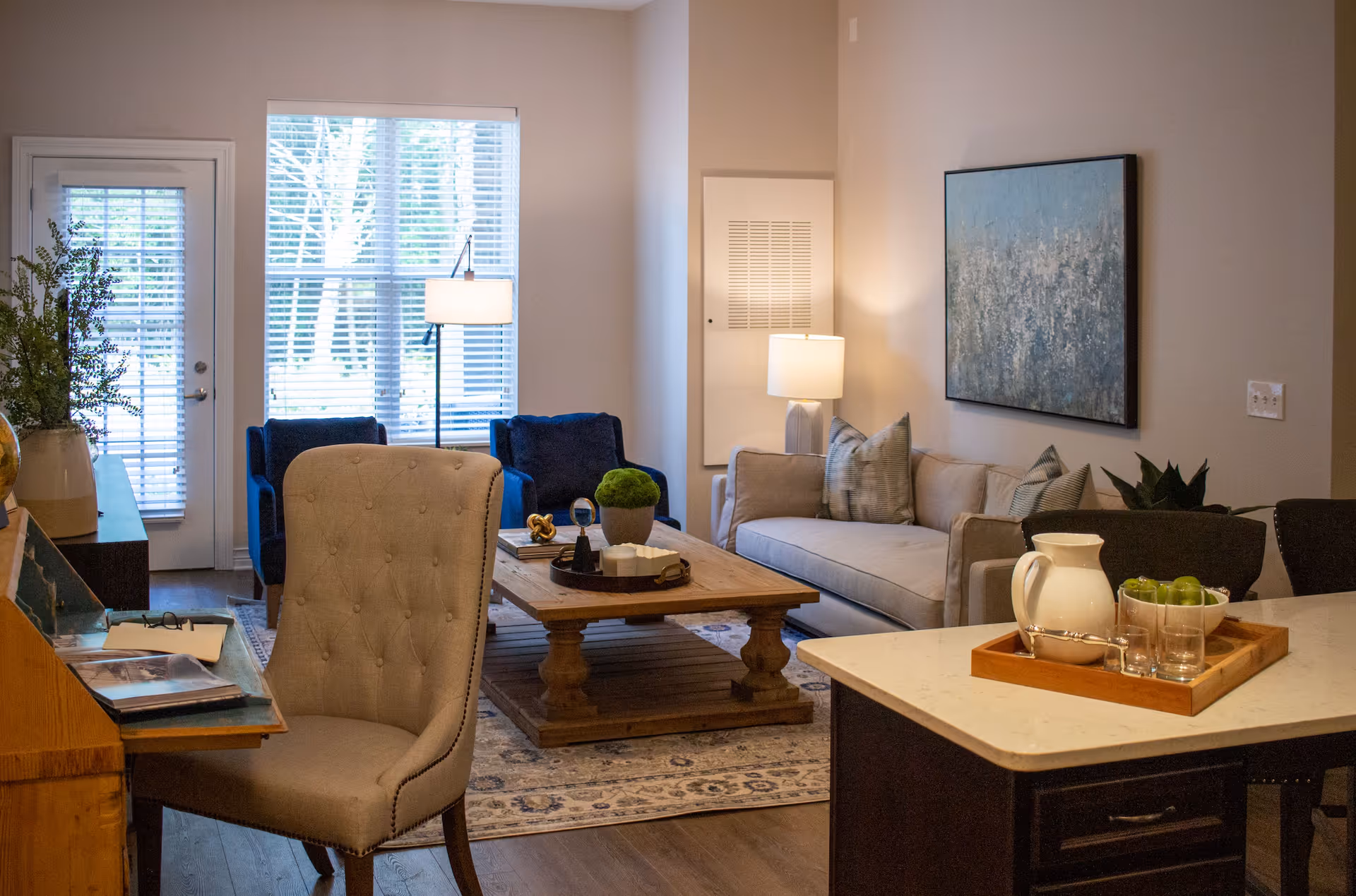 A cozy living room area with a beige tufted chair in the foreground, a wooden coffee table with decorative items, a beige sofa with patterned pillows, two blue armchairs, a floor lamp, and a large window with blinds letting in natural light. A kitchen island with a tray holding a white pitcher, glasses, and a bowl of green apples is visible on the right side.