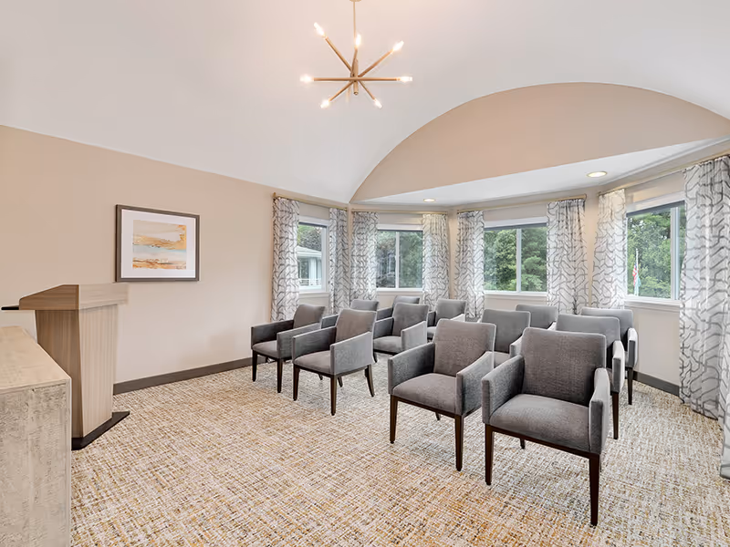 A small meeting or presentation room with two rows of gray upholstered chairs facing a wooden podium. The room has beige walls, a patterned carpet, large windows with sheer patterned curtains, and a modern chandelier hanging from a vaulted ceiling.