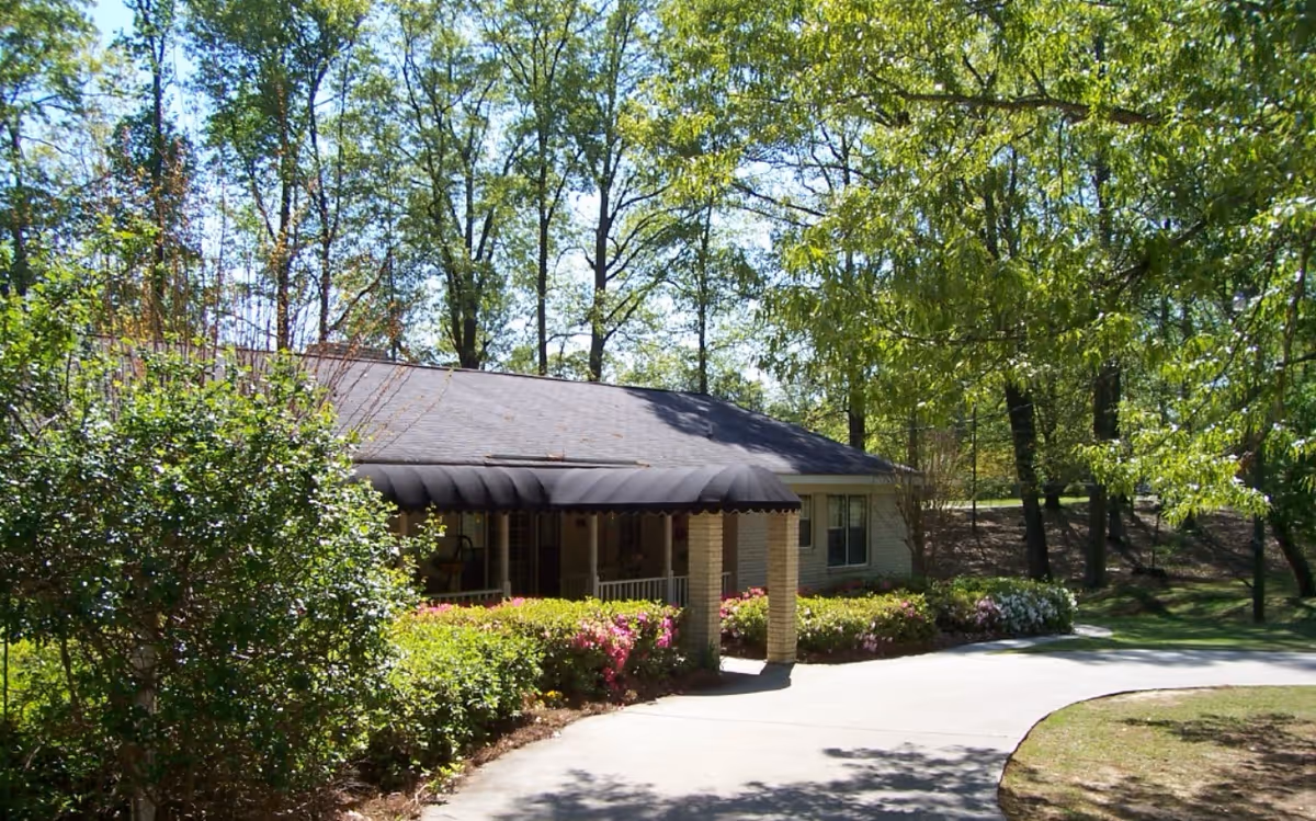 Front entrance of a single-story senior living building with a covered portico, driveway, and surrounding trees and shrubs.