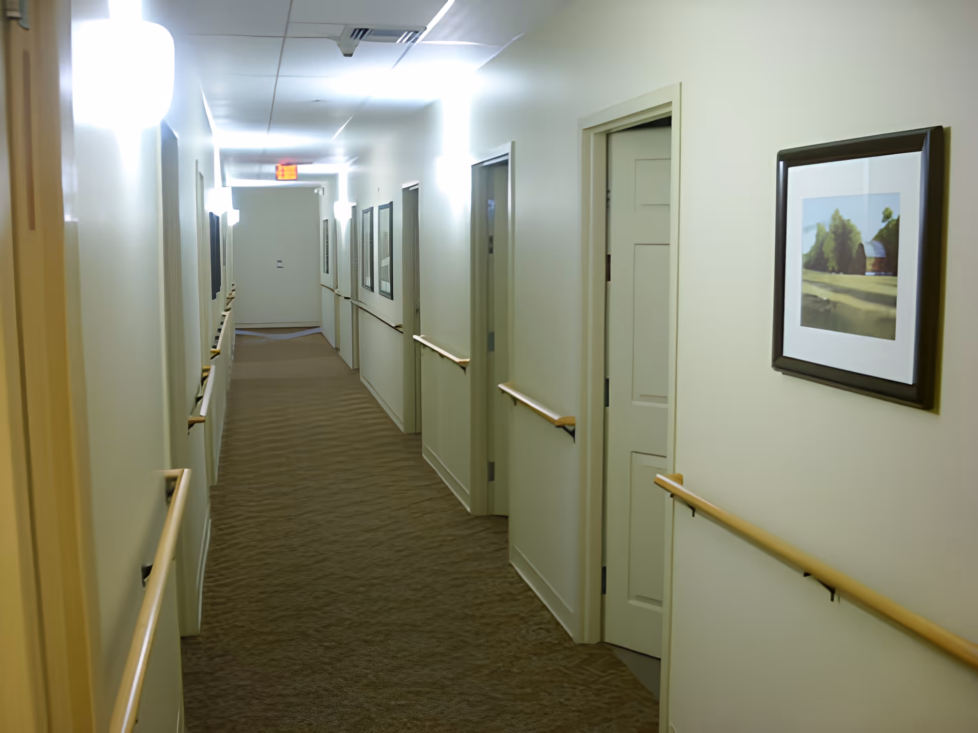 Well-lit interior hallway with handrails, multiple doors and framed artwork leading to an exit sign at the far end.