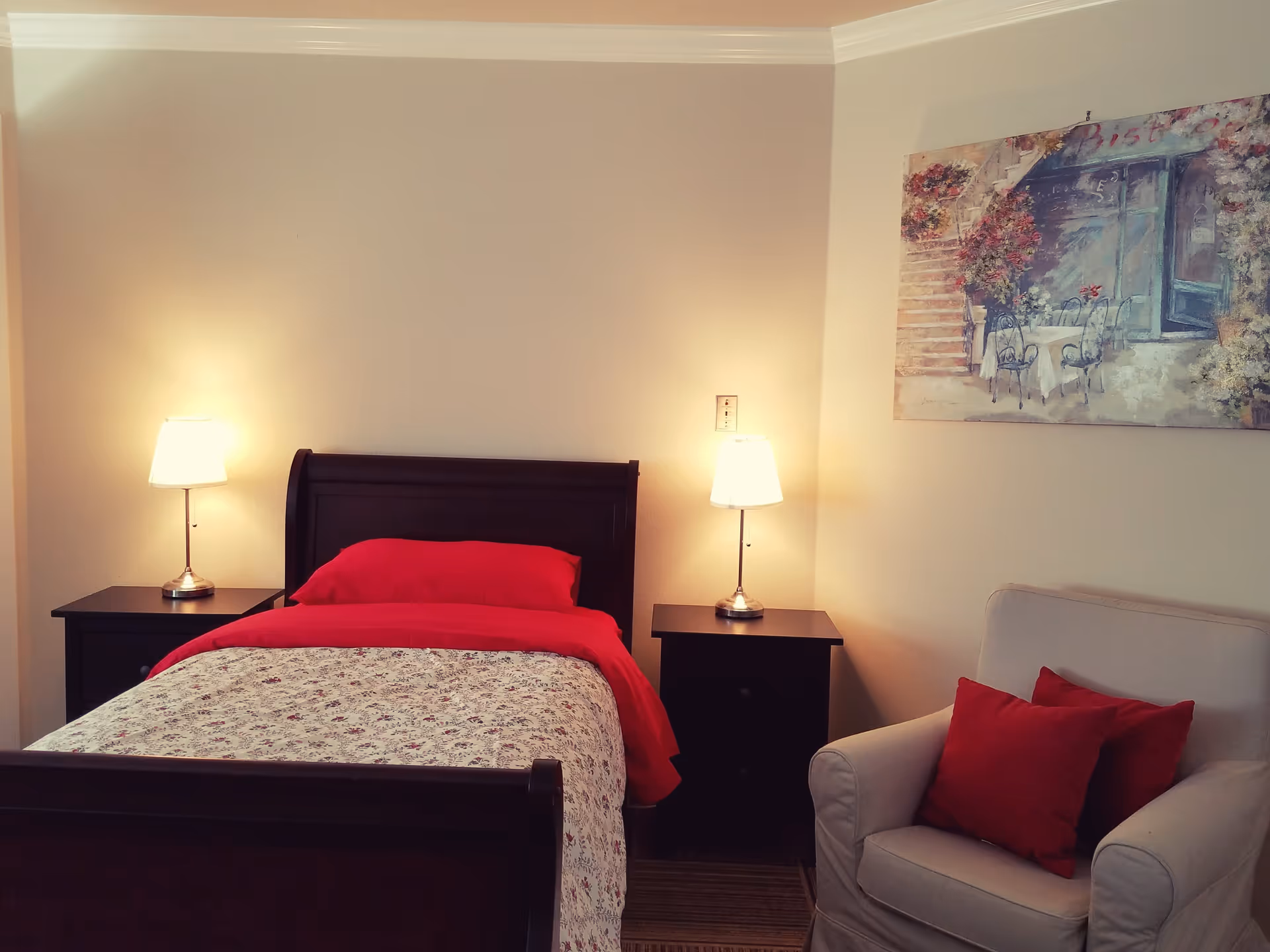 A cozy bedroom with a dark wooden bed frame, a bed with red pillow and blanket, and a floral patterned bedspread. There are two matching dark wooden nightstands on either side of the bed, each with a lit table lamp. To the right, there is a beige armchair with two red cushions. A colorful painting depicting an outdoor café scene hangs on the wall above the armchair.