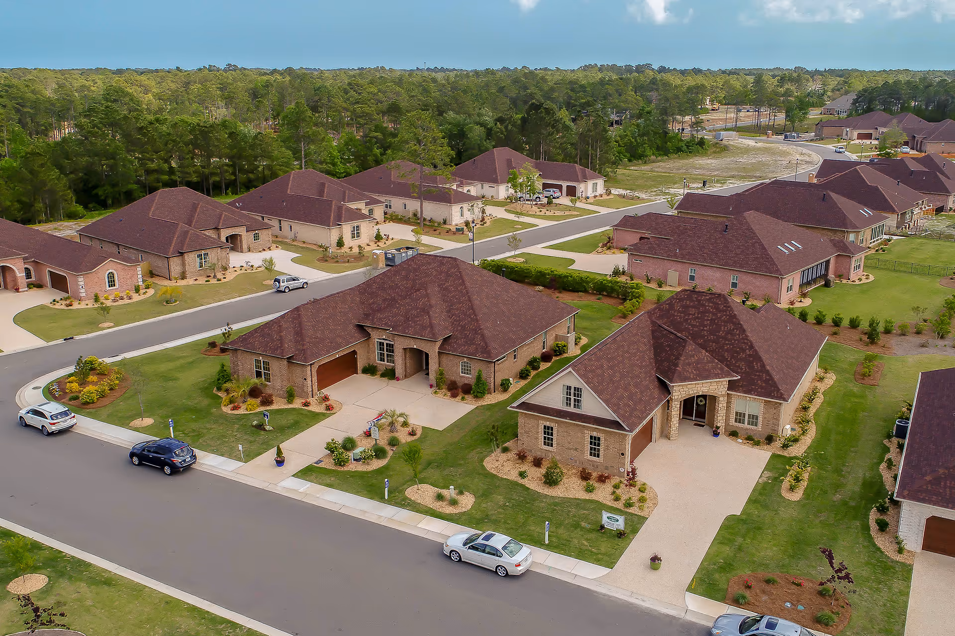 Aerial view of a residential neighborhood within The Village at Motts Landing, showing multiple single-story brick houses with brown roofs, well-maintained lawns, driveways, and several parked cars along the street. The area is surrounded by trees and greenery.
