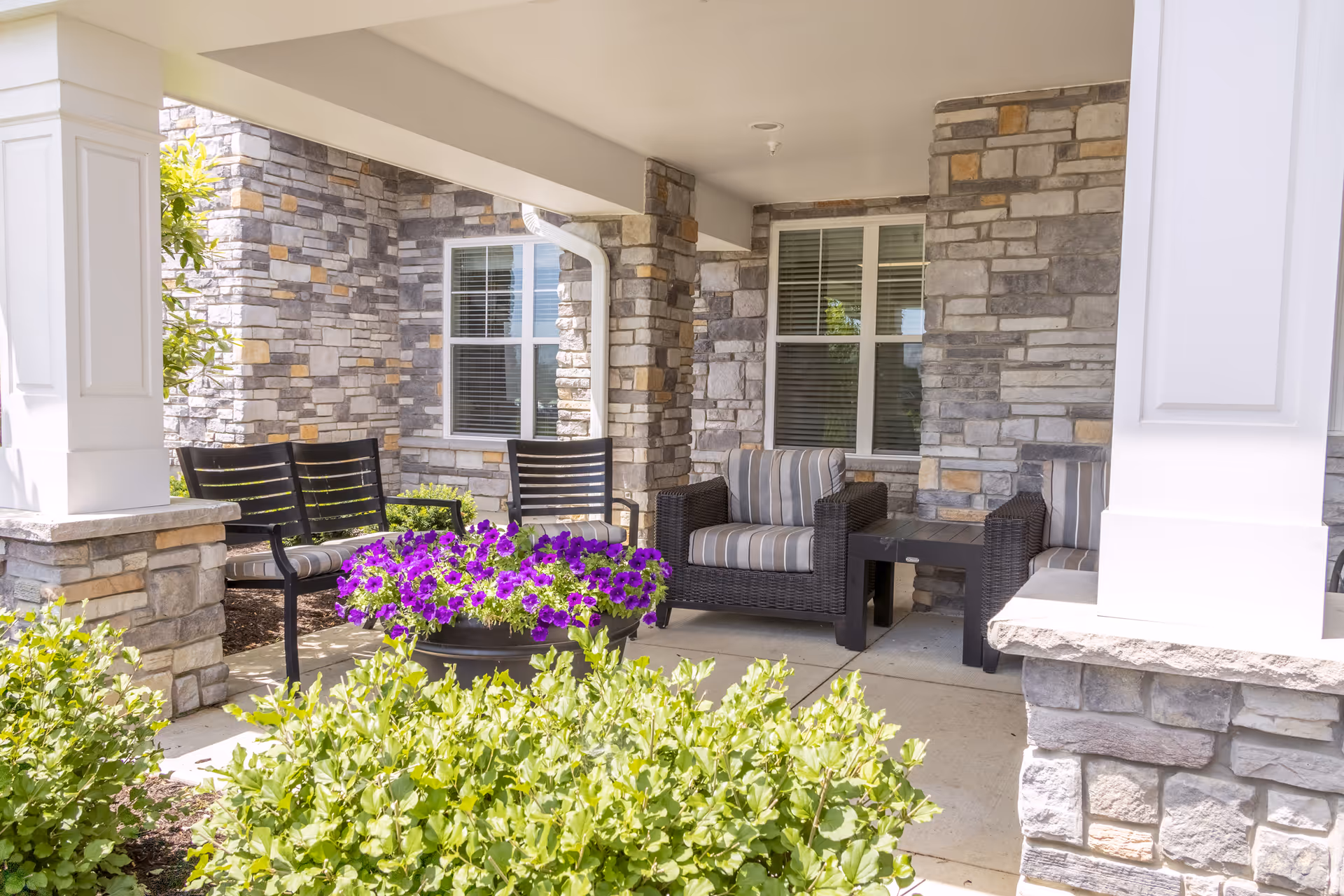 Covered outdoor seating area with stone pillars and walls, featuring two black chairs with striped cushions, a black side table, and a black bench. In front of the seating area, there is a planter with vibrant purple flowers and green shrubs surrounding the space.