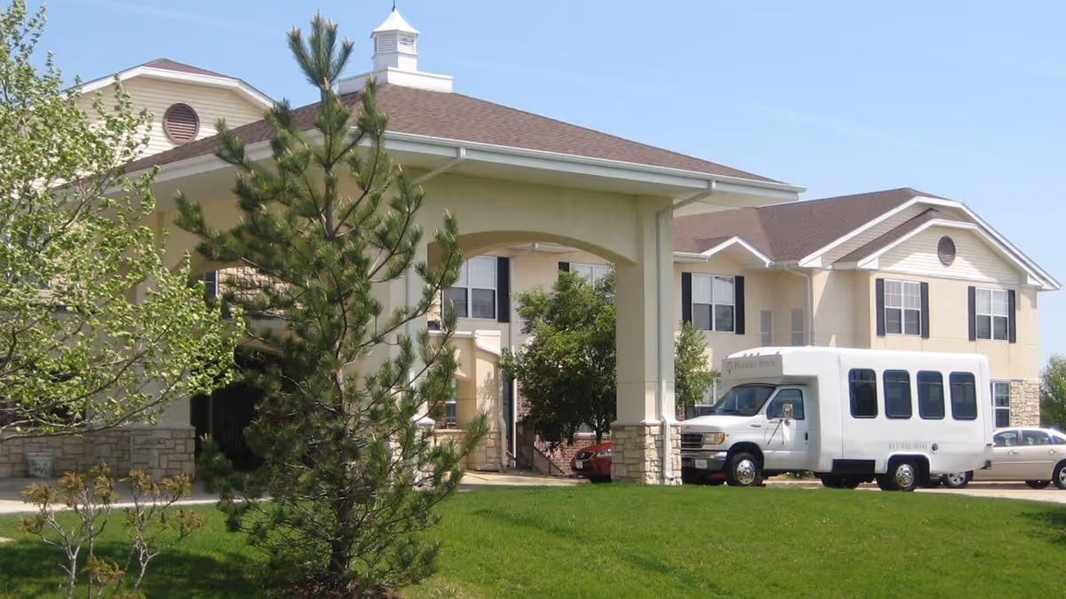 Exterior front entrance of a senior living facility with a covered porte-cochere, a shuttle van parked nearby, and a grassy lawn with trees.
