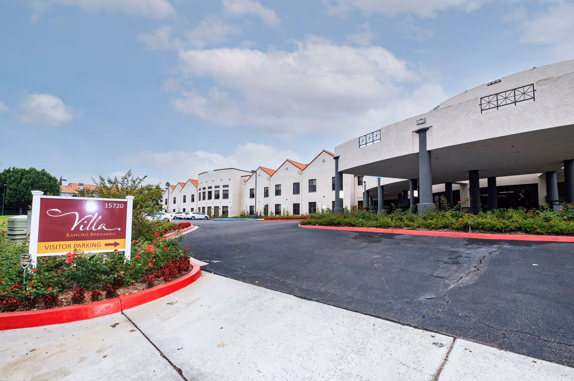 Exterior view of Villa Rancho Bernardo Care Center showing the main building with white walls and red-tiled roofs, a driveway, and a sign indicating visitor parking surrounded by landscaping with red flowers.