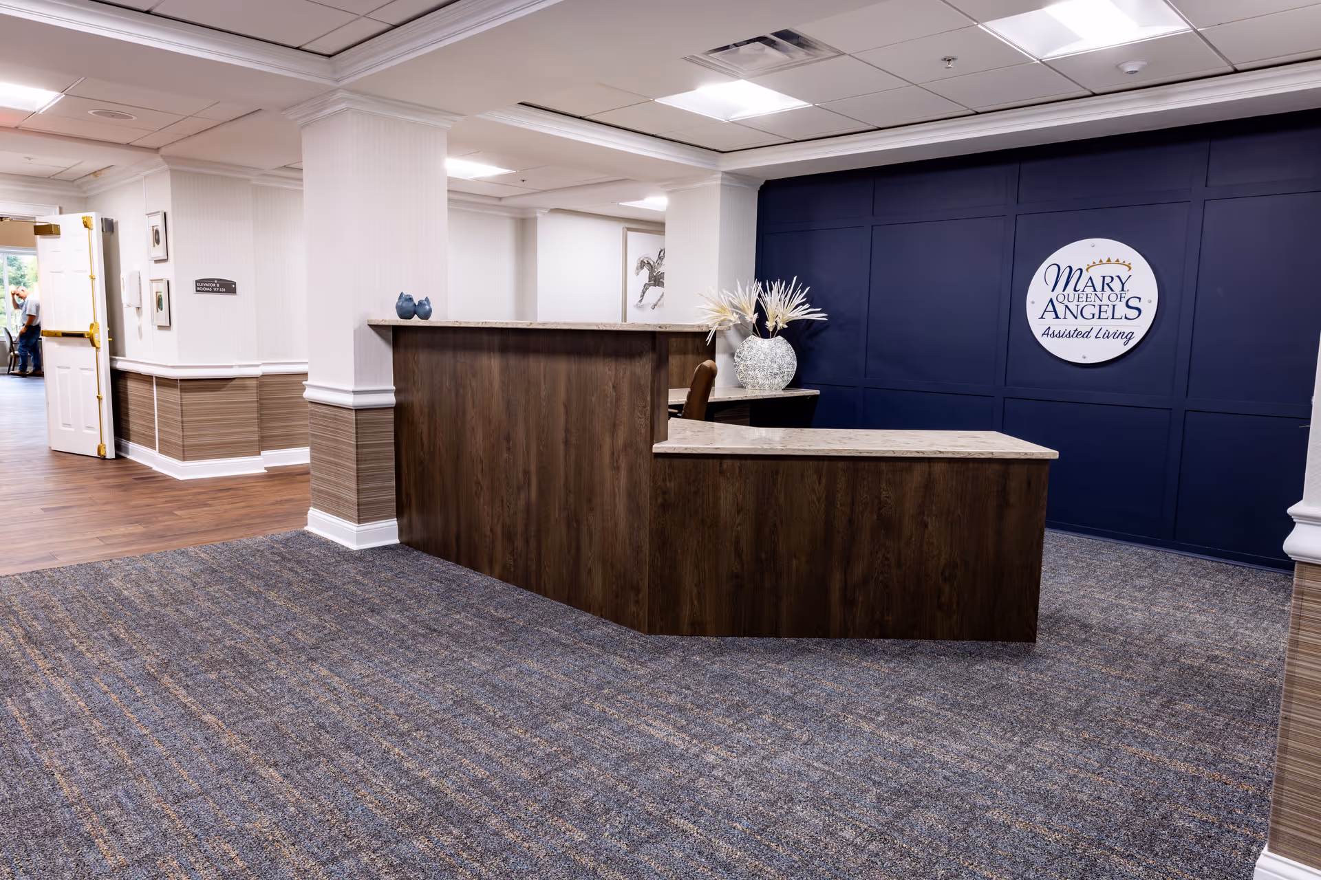 Reception desk and lobby area with a dark wood front desk, a blue feature wall displaying the Mary Queen of Angels Assisted Living logo, and carpeted flooring.