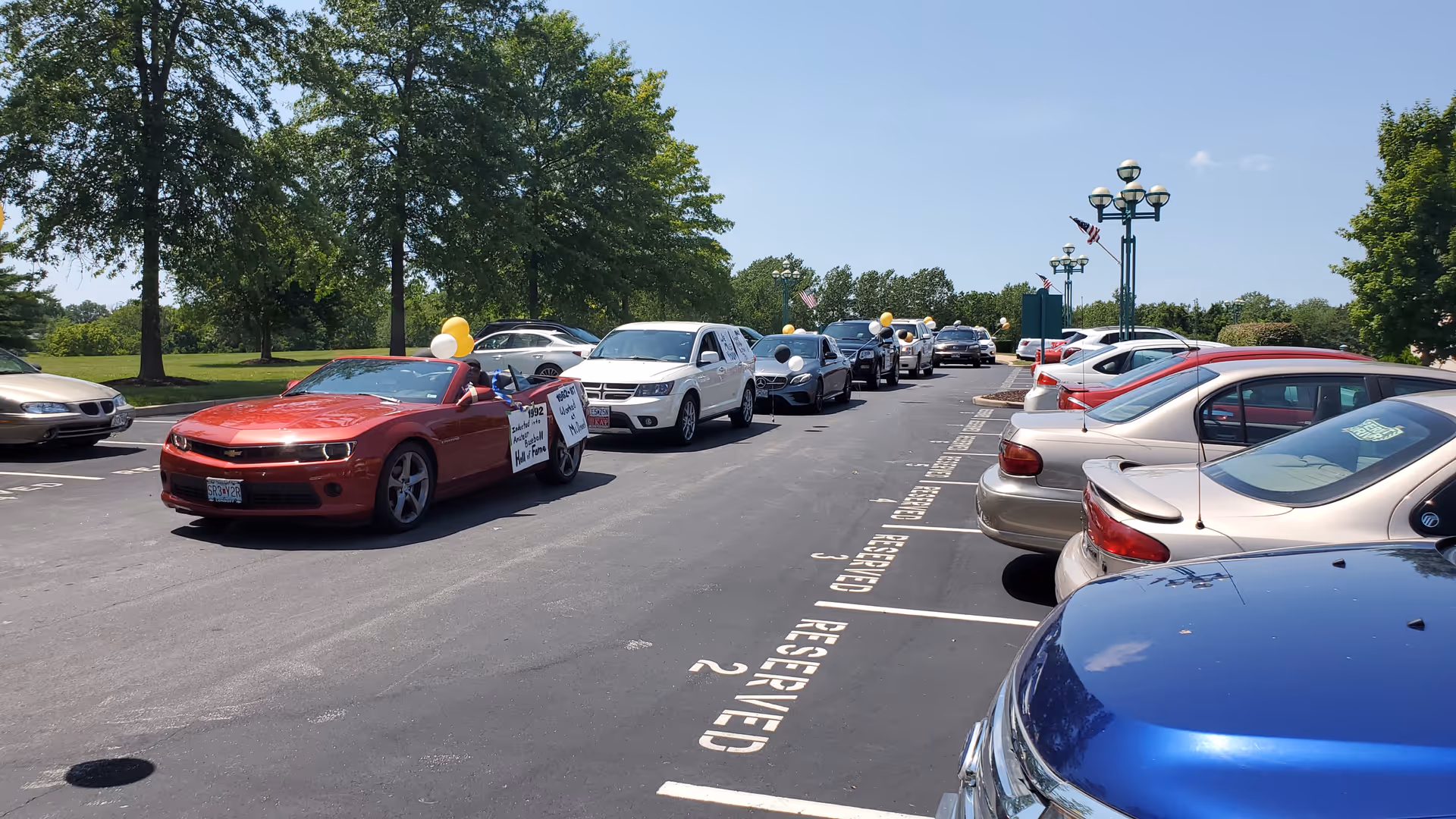 A parking lot with several parked cars on the right and a line of cars driving on the left. The first car in the line is a red convertible with balloons and a sign on the door. Trees and lamp posts are visible in the background under a clear blue sky.