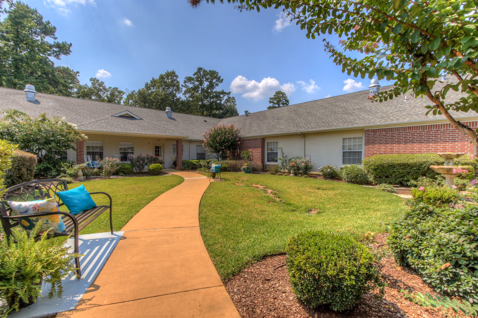 Sunny landscaped courtyard with a curved walkway, benches with colorful cushions, and a single-story senior living building surrounding it.
