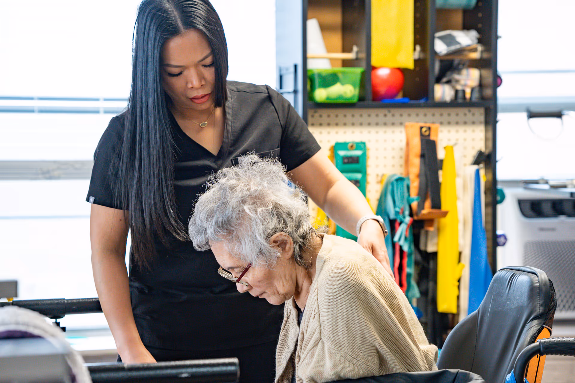 A caregiver in black scrubs assists an elderly woman with gray hair and glasses who is seated in a wheelchair. The background shows various colorful therapy or exercise equipment on shelves and hooks.