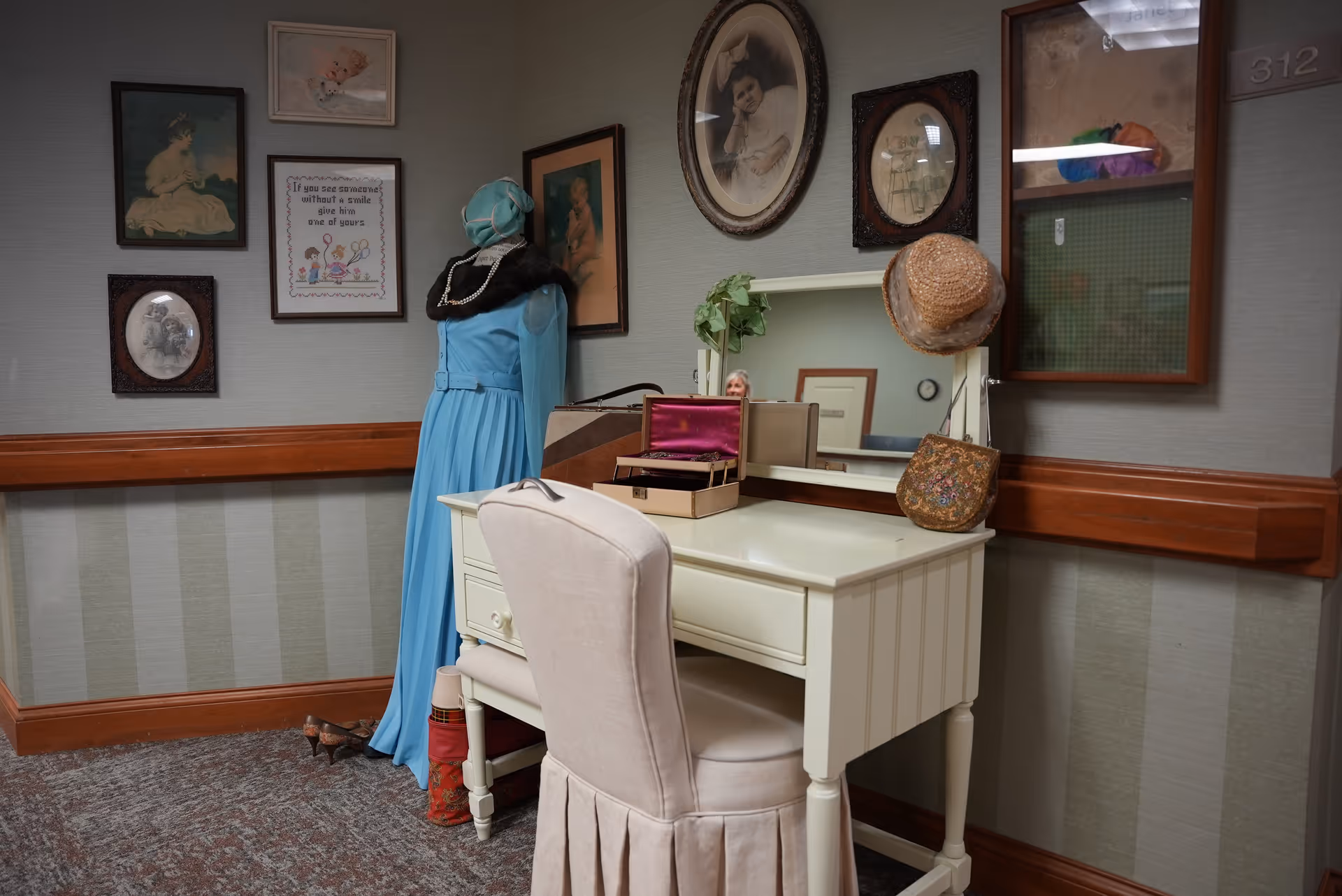A vintage-style vanity table with a mirror, a cushioned chair, and various decorative items including a hat and a jewelry box. Next to the vanity is a mannequin dressed in a long blue dress and a black shawl. The wall behind features several framed pictures and artwork. The setting appears to be a quiet, decorated corner of an indoor facility.
