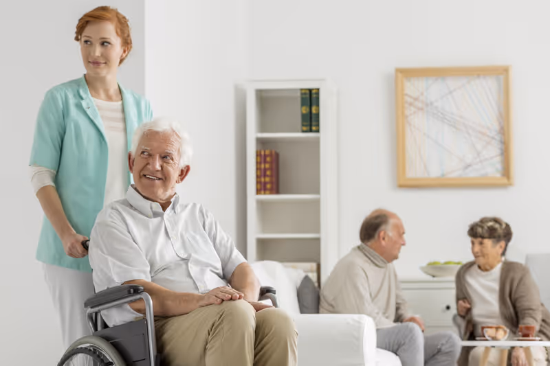 A caregiver stands behind an elderly man in a wheelchair, smiling. In the background, an elderly man and woman are seated on a couch, engaged in conversation in a bright, modern living room with a bookshelf and framed abstract artwork on the wall.