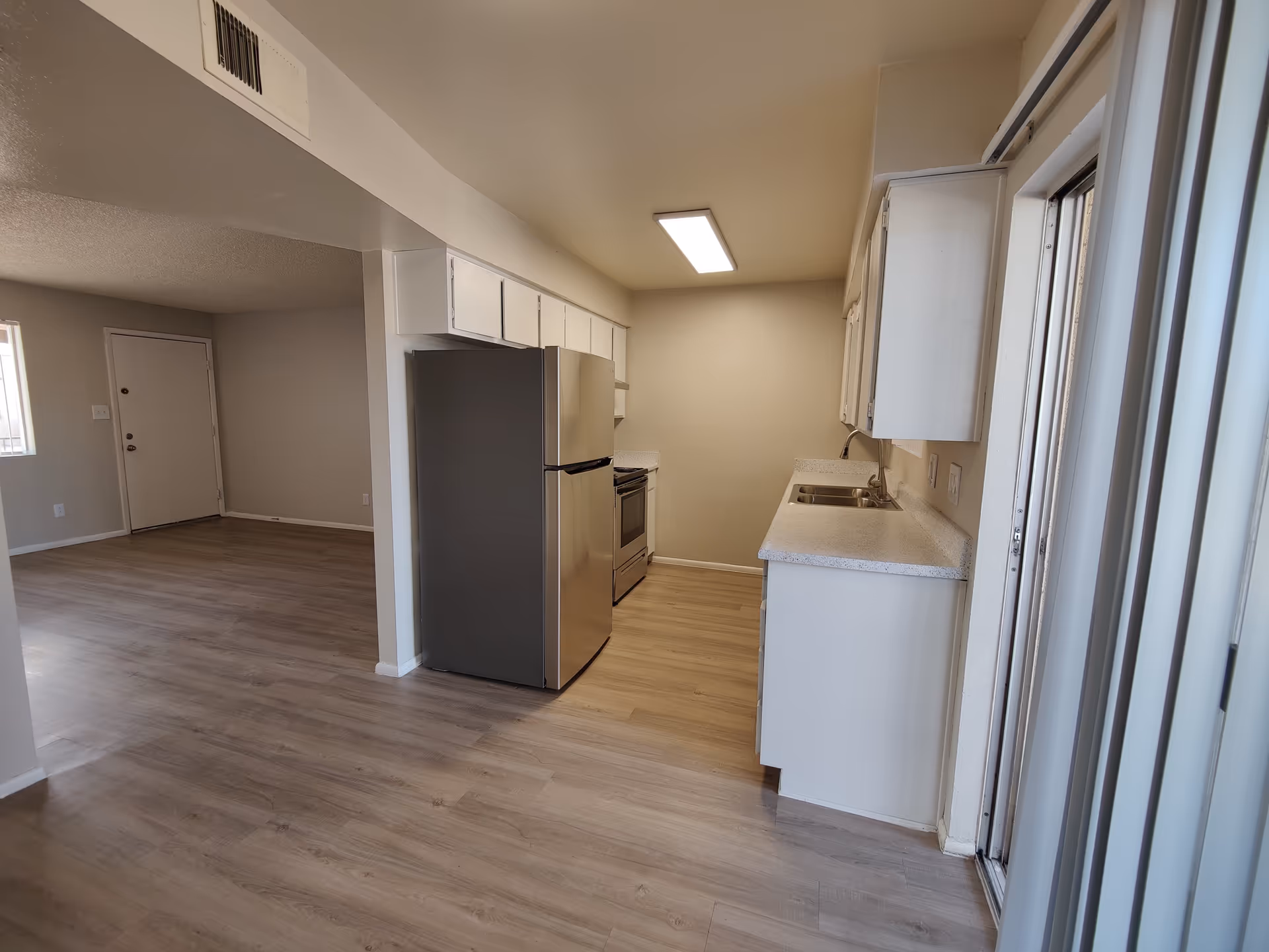 Interior view of an apartment kitchen with light wood flooring, white cabinets, a stainless steel refrigerator, stove, and a double sink. The kitchen opens into a living area with a door and window visible. Vertical blinds cover a sliding glass door on the right side.