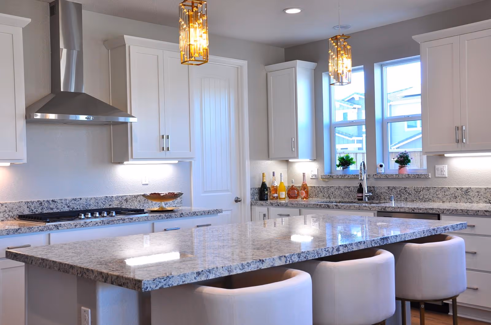 Bright modern kitchen with a large granite island, three upholstered stools, white cabinets, stainless range hood and pendant lights.