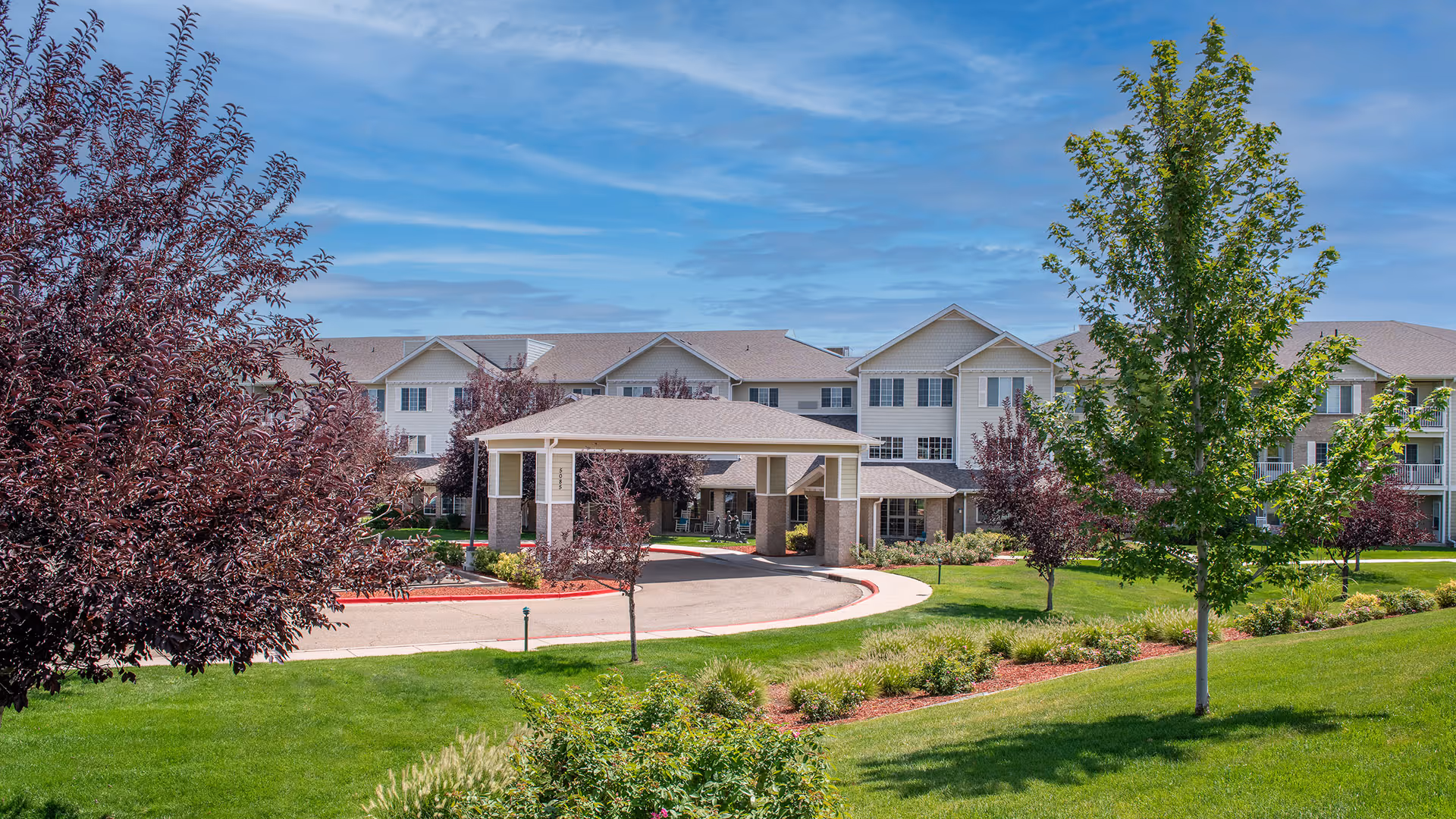 Multi-story senior living building exterior with a covered entrance, circular driveway, and landscaped lawn under a blue sky.