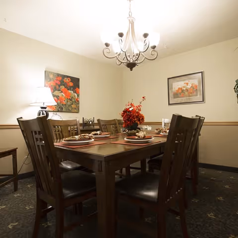 A dining room with a rectangular wooden table set for six people. The table has place settings with plates and napkins, and a centerpiece with red flowers. The room has beige walls with two floral paintings hanging, a table lamp on a side table, and a chandelier with five lights hanging from the ceiling.