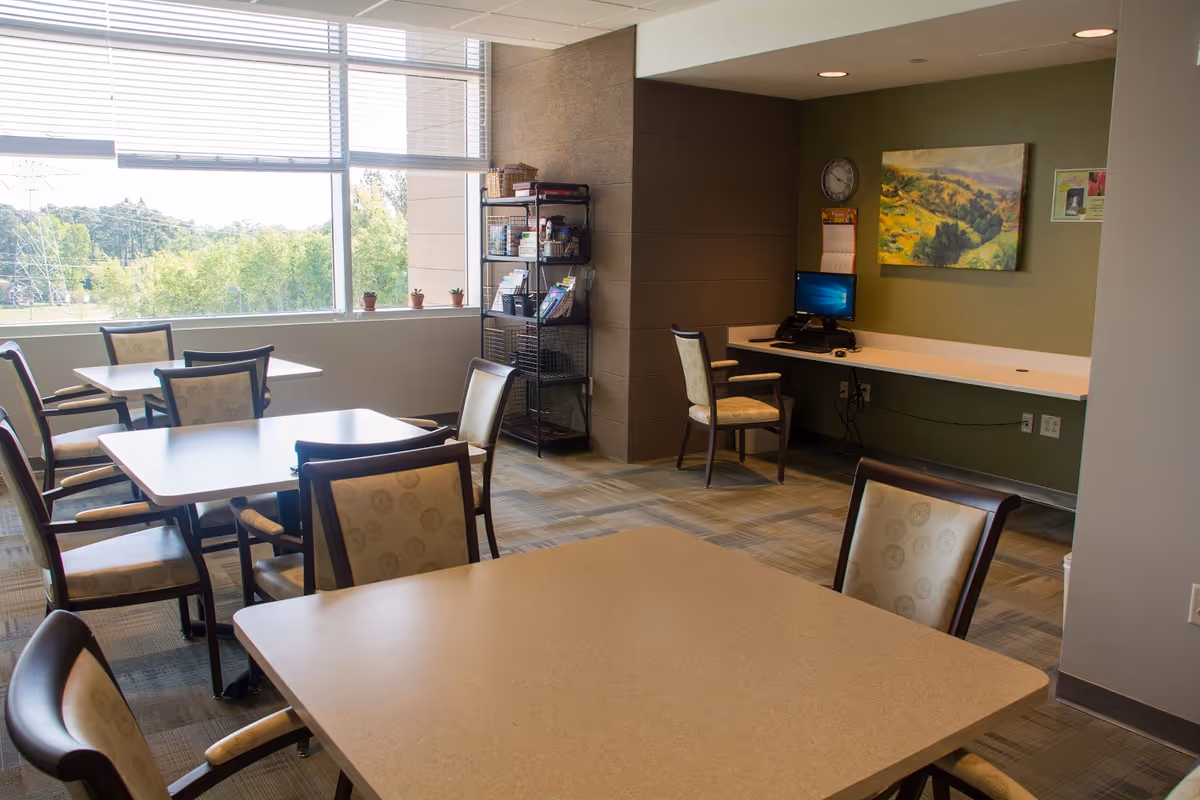 Bright communal dining/activity room with several tables and chairs, a computer workstation and shelving by a large window overlooking trees.