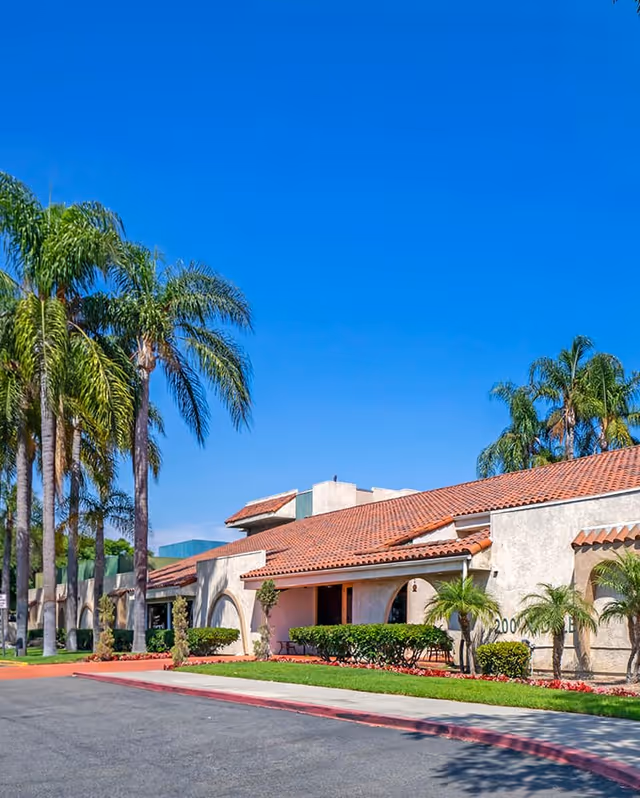 Exterior view of a single-story building with a red tile roof and beige stucco walls, surrounded by palm trees and neatly trimmed bushes under a clear blue sky.