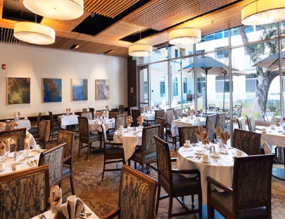 A bright dining room with multiple round and rectangular tables covered with white tablecloths, set with cups, plates, silverware, and folded napkins. The room features large floor-to-ceiling windows that look out onto an outdoor patio with umbrellas and trees. The ceiling has wooden slats and modern circular light fixtures, and there are abstract paintings on the wall.