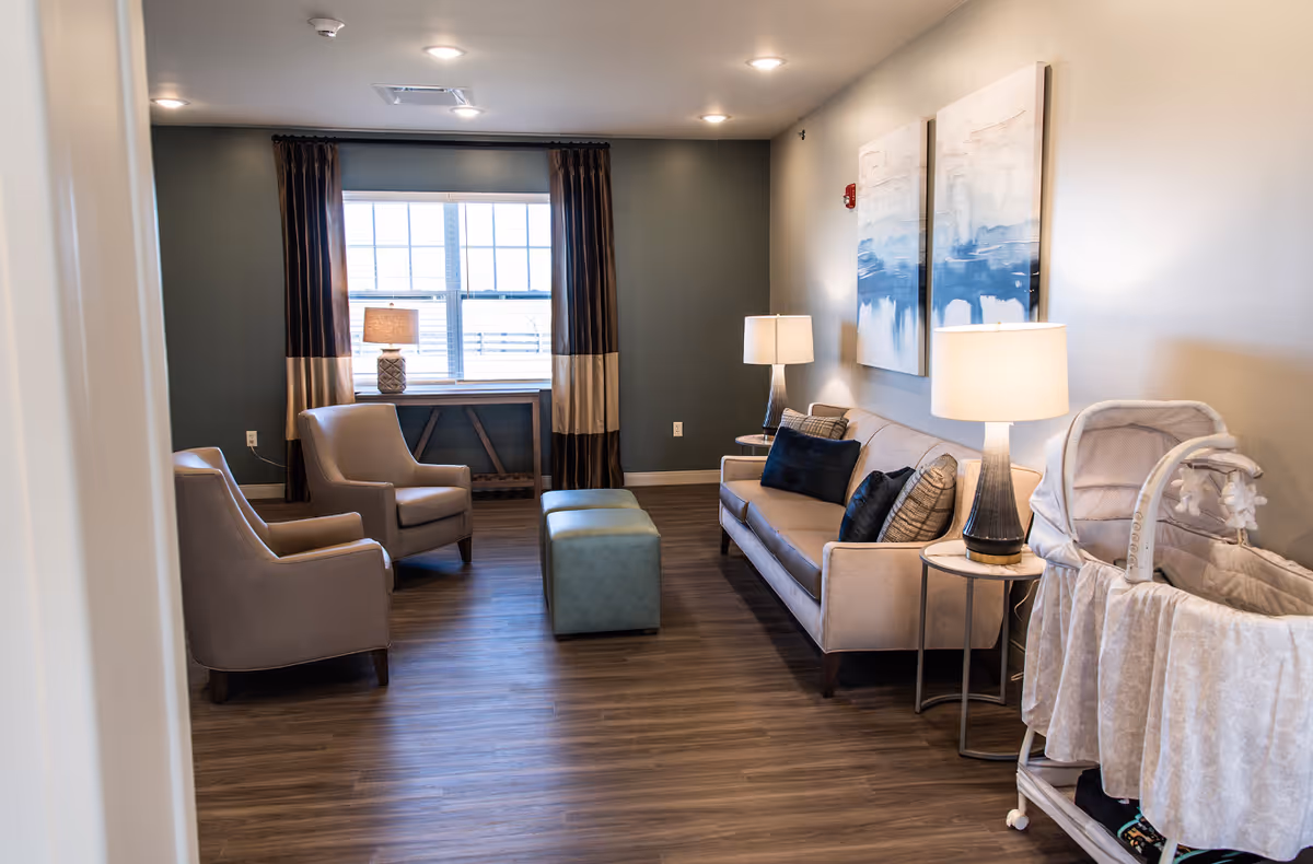 A cozy living room with two beige armchairs, a beige sofa with dark and patterned cushions, two side tables with lamps, a green ottoman, a window with curtains, and a baby bassinet on wheels. The walls are painted in neutral tones with two abstract paintings hanging above the sofa.