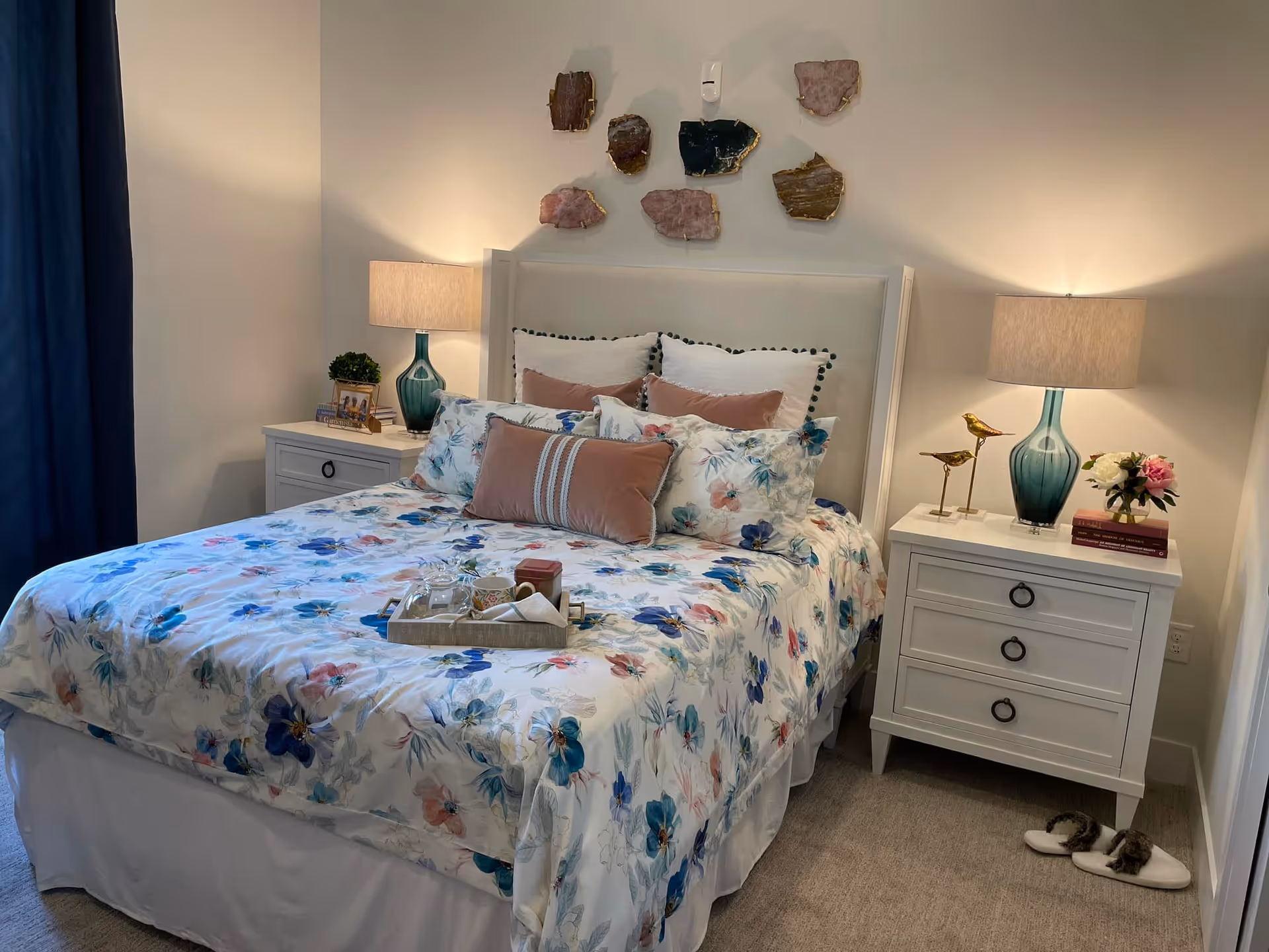 A neatly made bed with floral bedding and multiple pillows in a bedroom. There are two white nightstands on either side of the bed, each with a blue glass lamp. The left nightstand has a small plant and a framed photo, while the right nightstand has two decorative golden bird figurines, a small bouquet of flowers, and some books. A pair of slippers is placed on the carpeted floor next to the right nightstand. The wall above the bed features decorative stone-like wall art pieces.